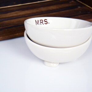May include: Two white ceramic bowls stacked on top of each other. The top bowl has the word "MRS." written in brown lettering.