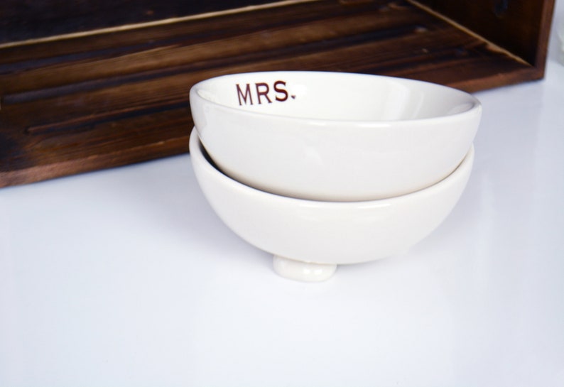 May include: Two white ceramic bowls stacked on top of each other. The top bowl has the word "MRS." written in brown lettering.