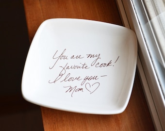 May include: A square white ceramic dish with rounded corners featuring dark brown cursive text that reads: You are my favorite cook! I love you - Mom, followed by a small hand-drawn heart symbol centered on the smooth, glossy surface.