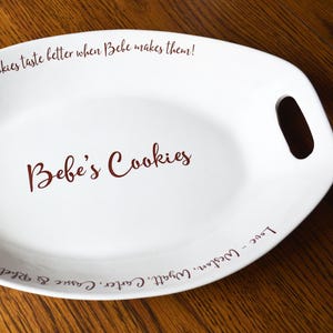 May include: White oval ceramic serving dish with handles. The dish has a red inscription that reads "Cookies taste better when Bebe makes them! Bebe's Cookies Love - Weston, Wyatt, Carter, Cassie & Rhett"