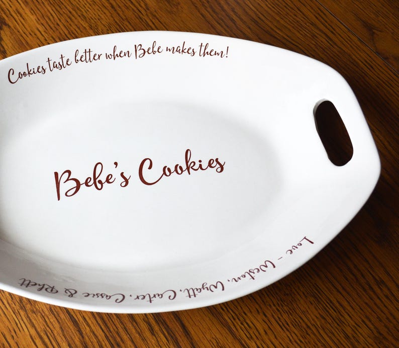 May include: White oval ceramic serving dish with handles. The dish has a red inscription that reads "Cookies taste better when Bebe makes them! Bebe's Cookies Love - Weston, Wyatt, Carter, Cassie & Rhett"