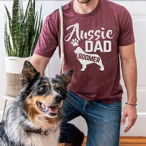 May include: A man wearing a maroon t-shirt with the text "Aussie Dad Boomer" and a silhouette of an Australian Shepherd dog. He is kneeling on a wooden floor with a blue merle Australian Shepherd dog sitting beside him.