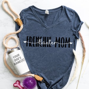 Op de afbeelding: Een marineblauw V-hals T-shirt met de tekst "FRENCHIE MOM" en de naam "Baxter" in witte letters. Een witte beker met "YOUR DOG'S NAME(S)", een beige touwriem en een paars hondenspeeltje zijn ook zichtbaar.