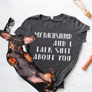 Puede incluir: Una camiseta gris con el texto "My Dachshund and I Talk Shit About You" en ella. Un dachshund negro est&aacute; acostado sobre la camiseta.