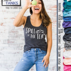 May include: A woman wearing a gray tank top with the text "Spill the Tea" and two teacup illustrations. The tank top is available in a variety of colors, including red pink, peach, white, gray, charcoal, navy, purple, royal, turquoise, and teal.