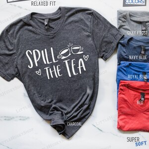 May include: A charcoal gray V-neck t-shirt with the text "Spill the Tea" in white. The shirt has a relaxed fit and is available in other colors, including gray frost, navy blue, royal blue, and red.