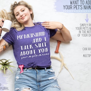 Puede incluir: Camiseta morada con el texto "My Dachshund and I Talk Shit About You" con un espacio para el nombre de la mascota. El texto est&aacute; en blanco. La modelo lleva la camiseta y est&aacute; tumbada sobre una superficie blanca.