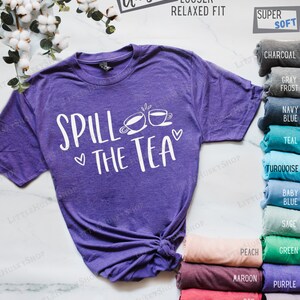 May include: Purple t-shirt with the text "Spill the Tea" and two teacups. The shirt is available in a variety of colors, including charcoal, gray frost, navy blue, teal, turquoise, baby blue, sage, peach, green, maroon, purple, red, royal blue, and lavender.