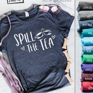 May include: A navy blue t-shirt with the text "Spill the Tea" and two teacups. The shirt is semi-fitted and runs small. The shirt is available in a variety of colors, including charcoal, gray frost, navy blue, teal, turquoise, baby blue, green, purple, maroon, red, and royal blue.