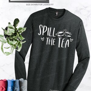 May include: A charcoal grey long-sleeve t-shirt with the text "Spill the Tea" in white lettering and two white teacup illustrations. The shirt is a unisex relaxed fit.