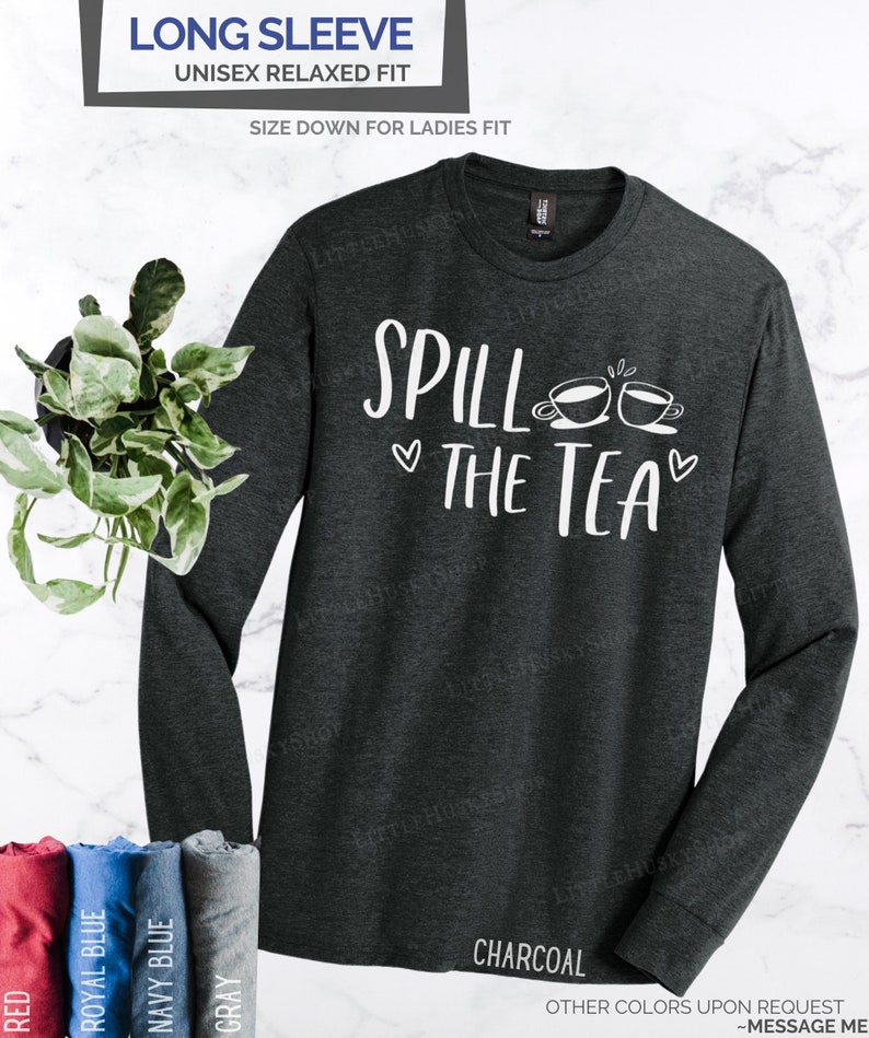 May include: A charcoal grey long-sleeve t-shirt with the text "Spill the Tea" in white lettering and two white teacup illustrations. The shirt is a unisex relaxed fit.