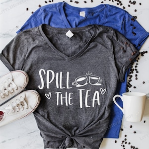 May include: A dark gray V-neck t-shirt with the text "Spill the Tea" in white lettering. The text is accompanied by a graphic of two teacups with steam coming from them. The shirt is tied in a knot at the bottom.