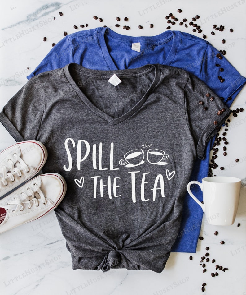 May include: A dark gray V-neck t-shirt with the text "Spill the Tea" in white lettering. The text is accompanied by a graphic of two teacups with steam coming from them. The shirt is tied in a knot at the bottom.