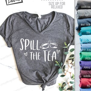 May include: A heather gray V-neck t-shirt with the text "Spill the Tea" in white lettering. Two white teacup illustrations are above the text. The shirt is semi-fitted and runs small. The text "Super Soft" is in a white box in the upper right corner of the image. The image shows a variety of colors for the shirt, including charcoal, gray frost, navy blue, teal, turquoise, baby blue, green, purple, maroon, red, royal blue, and peach.