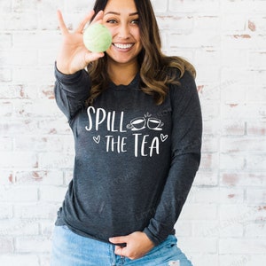 May include: A woman wearing a dark grey long-sleeved shirt with the text "Spill the Tea" and two teacup icons. She is holding a green tennis ball in her right hand.