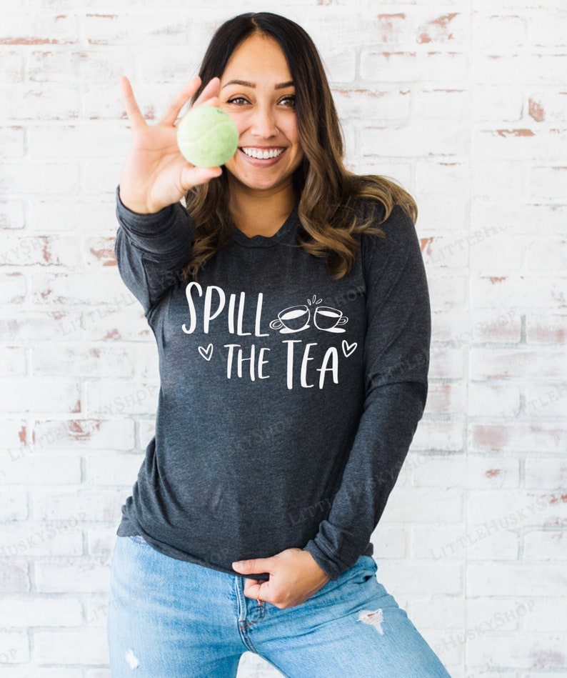 May include: A woman wearing a dark grey long-sleeved shirt with the text "Spill the Tea" and two teacup icons. She is holding a green tennis ball in her right hand.