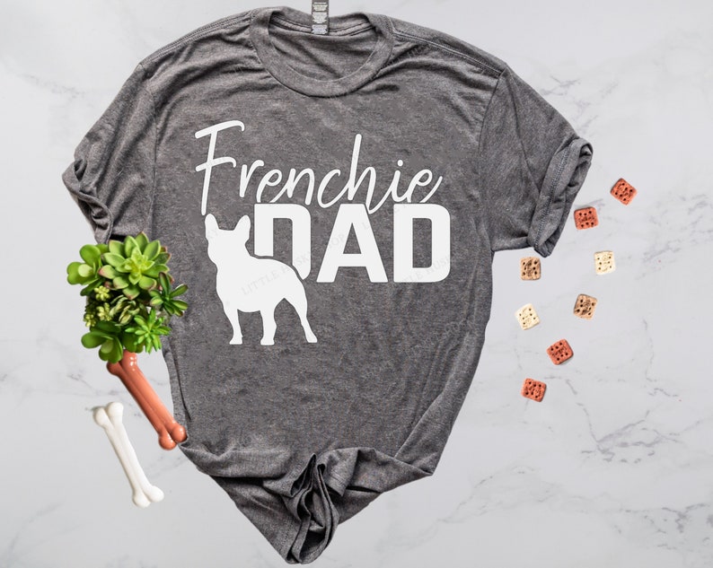 May include: A heather gray t-shirt with the words "Frenchie DAD" in white, along with a white silhouette of a French Bulldog. The shirt is styled with dog treats and a small succulent plant.