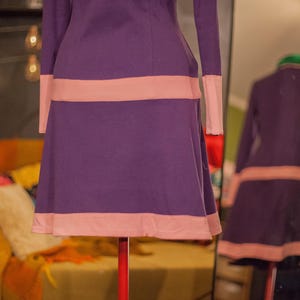 May include: A long-sleeved dress in a deep purple hue featuring horizontal light pink bands across the waist and the hemline. The garment has a fitted bodice that transitions into a flared skirt, with light pink cuffs on the sleeves.