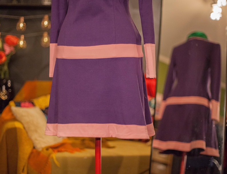 May include: A long-sleeved dress in a deep purple hue featuring horizontal light pink bands across the waist and the hemline. The garment has a fitted bodice that transitions into a flared skirt, with light pink cuffs on the sleeves.