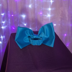 May include: A large turquoise satin bow with a gathered elastic band in the center. The bow is sitting on a dark purple surface.