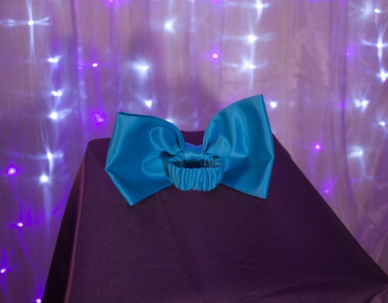 May include: A large turquoise satin bow with a gathered elastic band in the center. The bow is sitting on a dark purple surface.