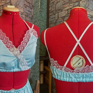 May include: Pale blue satin lingerie set with white lace detailing. The top has a wrap-style front and spaghetti straps. The back of the top has criss-cross straps. The set is displayed on a red dress form.