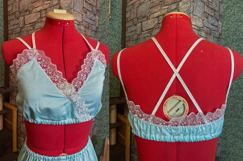 May include: Pale blue satin lingerie set with white lace detailing. The top has a wrap-style front and spaghetti straps. The back of the top has criss-cross straps. The set is displayed on a red dress form.