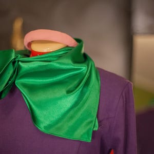 May include: A vibrant emerald green satin scarf tied around the neck of a mannequin wearing a solid purple long-sleeved top. A smooth, rounded light pink headband is positioned on the head of the mannequin above the scarf.