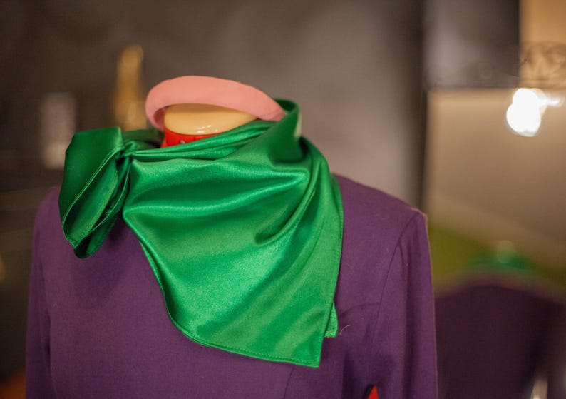 May include: A vibrant emerald green satin scarf tied around the neck of a mannequin wearing a solid purple long-sleeved top. A smooth, rounded light pink headband is positioned on the head of the mannequin above the scarf.
