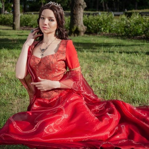 May include: A red medieval-style dress with long, flowing sleeves and a full skirt. The bodice is a darker shade of red with floral embroidery. The dress is worn with a silver tiara and necklace. The model is sitting on green grass.