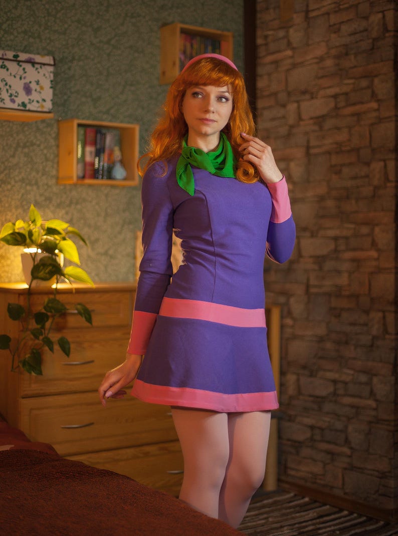 May include: A long-sleeved purple dress featuring a pink band at the waist and hem, paired with a bright green neck scarf and a matching pink headband. The dress has a fitted bodice and a flared skirt, worn with light-colored opaque tights.