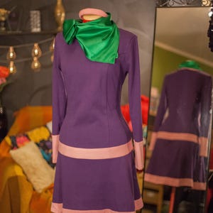 May include: A long-sleeved purple dress featuring a light pink horizontal band at the waist and hem, paired with a bright green satin neck scarf. The dress has a simple round neckline and a flared skirt silhouette, displayed on a mannequin.