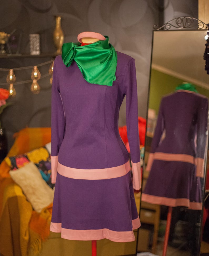 May include: A long-sleeved purple dress featuring a light pink horizontal band at the waist and hem, paired with a bright green satin neck scarf. The dress has a simple round neckline and a flared skirt silhouette, displayed on a mannequin.