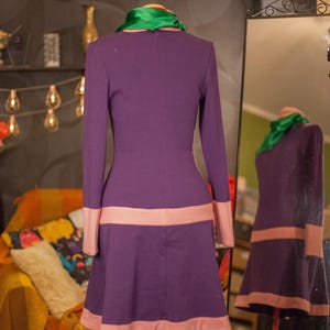 May include: A long-sleeved purple dress featuring a bright green scarf at the neckline and light pink horizontal bands at the waist and hemline. The dress is displayed on a mannequin, with a mirror reflection showing the back view.
