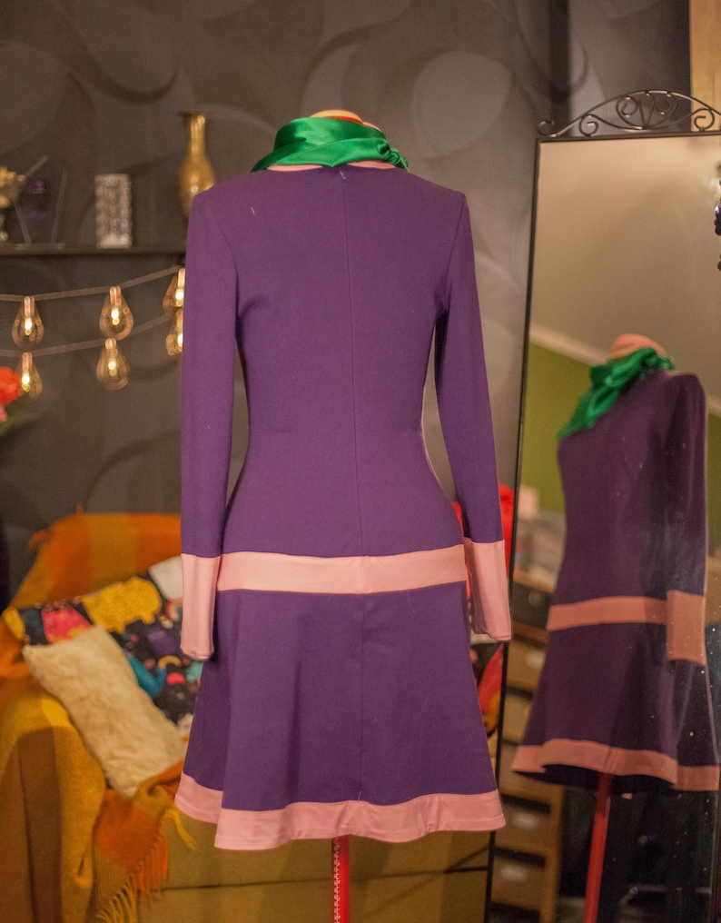 May include: A long-sleeved purple dress featuring a bright green scarf at the neckline and light pink horizontal bands at the waist and hemline. The dress is displayed on a mannequin, with a mirror reflection showing the back view.