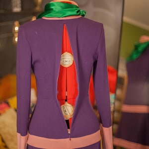 May include: A long-sleeved purple dress featuring a high neckline with a green scarf, a wide light pink horizontal band at the waist, and matching light pink cuffs. The back of the dress has a long vertical slit opening, revealing a red mannequin.