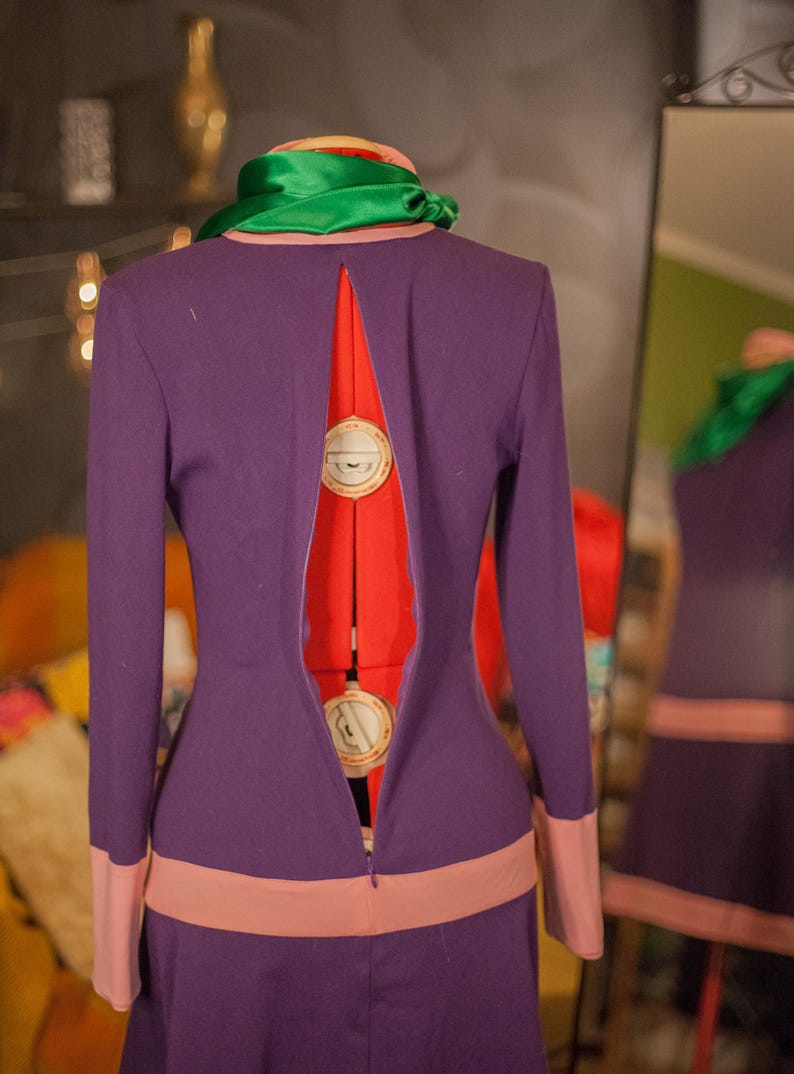 May include: A long-sleeved purple dress featuring a high neckline with a green scarf, a wide light pink horizontal band at the waist, and matching light pink cuffs. The back of the dress has a long vertical slit opening, revealing a red mannequin.