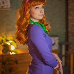 May include: A long-sleeved purple dress with a pink waistband, paired with a bright green neck scarf and a pink headband. The outfit features long, wavy orange hair and is designed to replicate a specific character costume.