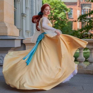 May include: A woman in a yellow gown with a blue sash and a crown on her head. She is twirling and her dress is flowing in the wind.