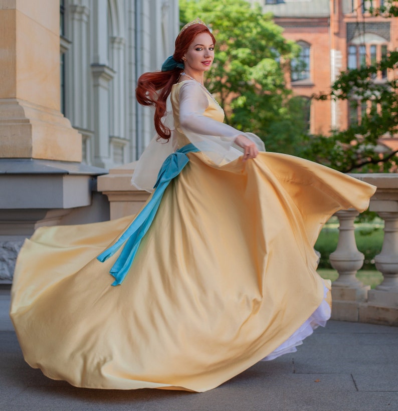 May include: A woman in a yellow gown with a blue sash and a crown on her head. She is twirling and her dress is flowing in the wind.