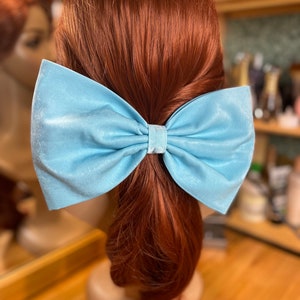 May include: A light blue satin bow hair clip. The bow is tied in a large, full loop with a center knot. The bow is attached to a hair clip.