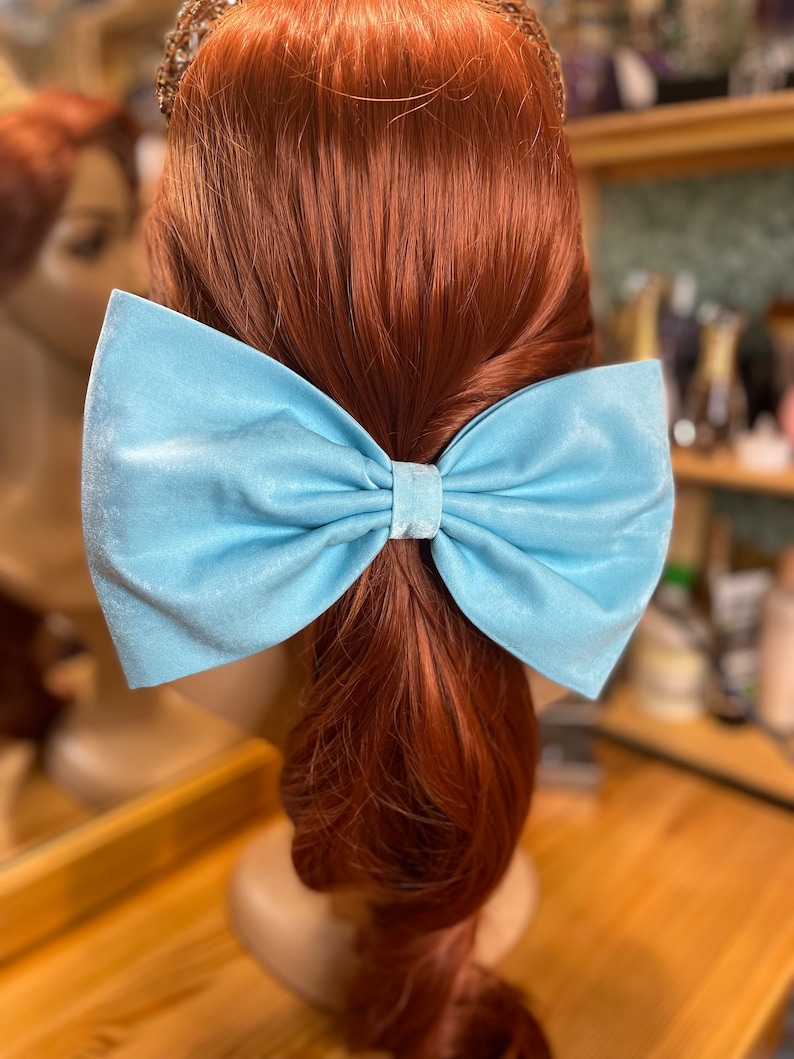 May include: A light blue satin bow hair clip. The bow is tied in a large, full loop with a center knot. The bow is attached to a hair clip.
