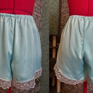 May include: Pale blue satin shorts with an elasticated waistband and white lace trim. The shorts are displayed on a red dress form. The garment is a vintage-inspired piece of lingerie.