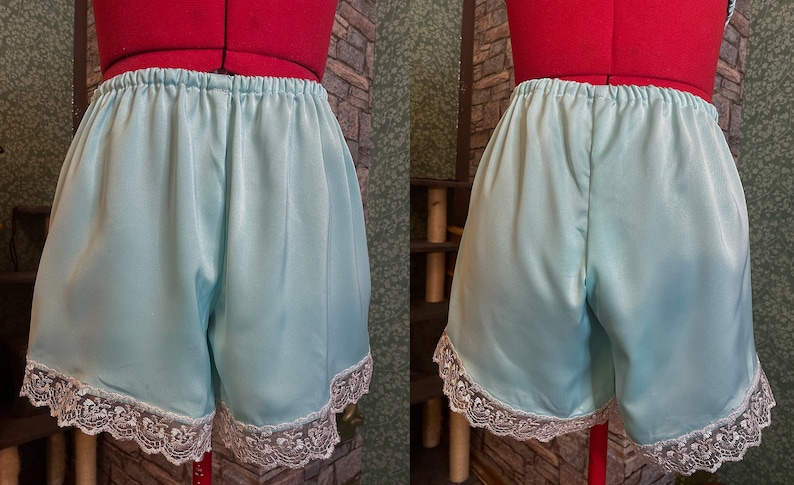 May include: Pale blue satin shorts with an elasticated waistband and white lace trim. The shorts are displayed on a red dress form. The garment is a vintage-inspired piece of lingerie.