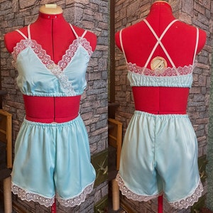 May include: A light blue satin lingerie set, comprising a crop top and shorts, displayed on a red dress form. The top has a wrap-style design with white lace trim. The shorts also feature white lace trim.