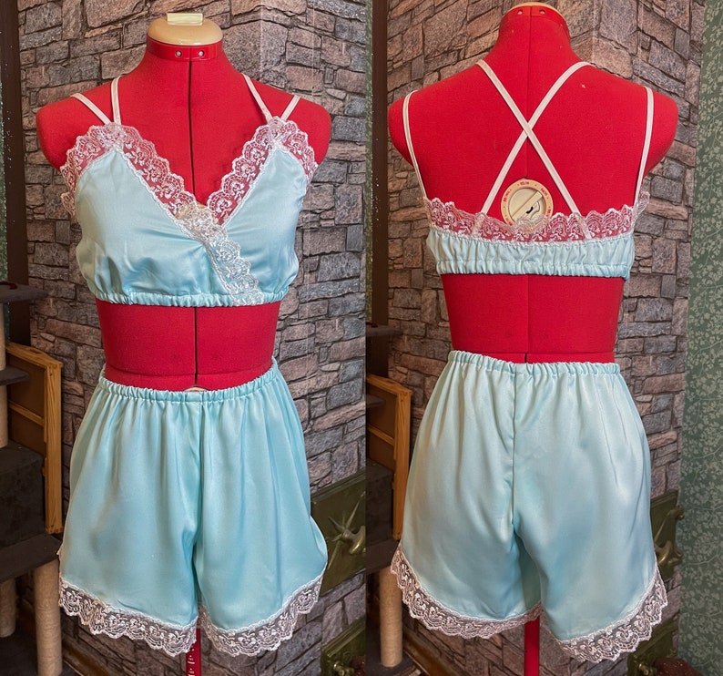 May include: A light blue satin lingerie set, comprising a crop top and shorts, displayed on a red dress form. The top has a wrap-style design with white lace trim. The shorts also feature white lace trim.