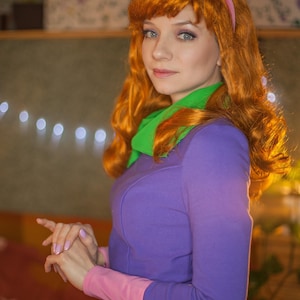 May include: A long-sleeved purple dress with a pink hem and cuffs, paired with a bright green neck scarf. The outfit includes a pink headband worn over long, wavy orange hair. The ensemble features a vibrant color palette and a fitted silhouette.