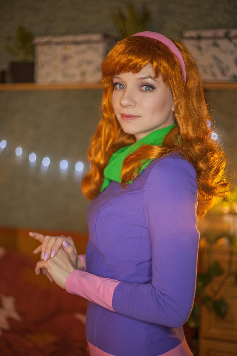 May include: A long-sleeved purple dress with a pink hem and cuffs, paired with a bright green neck scarf. The outfit includes a pink headband worn over long, wavy orange hair. The ensemble features a vibrant color palette and a fitted silhouette.