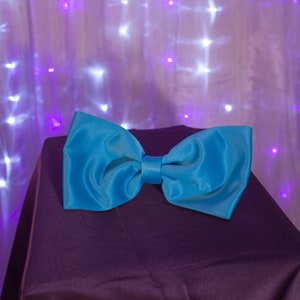 May include: A bright blue bow tie made of satin fabric, lying on a dark purple surface. The bow tie is a popular accessory for formal events.