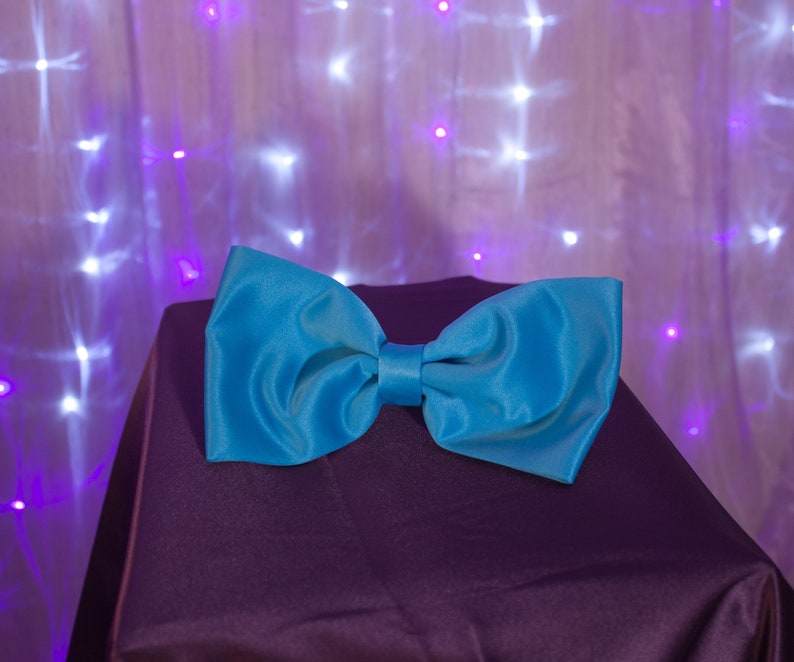 May include: A bright blue bow tie made of satin fabric, lying on a dark purple surface. The bow tie is a popular accessory for formal events.